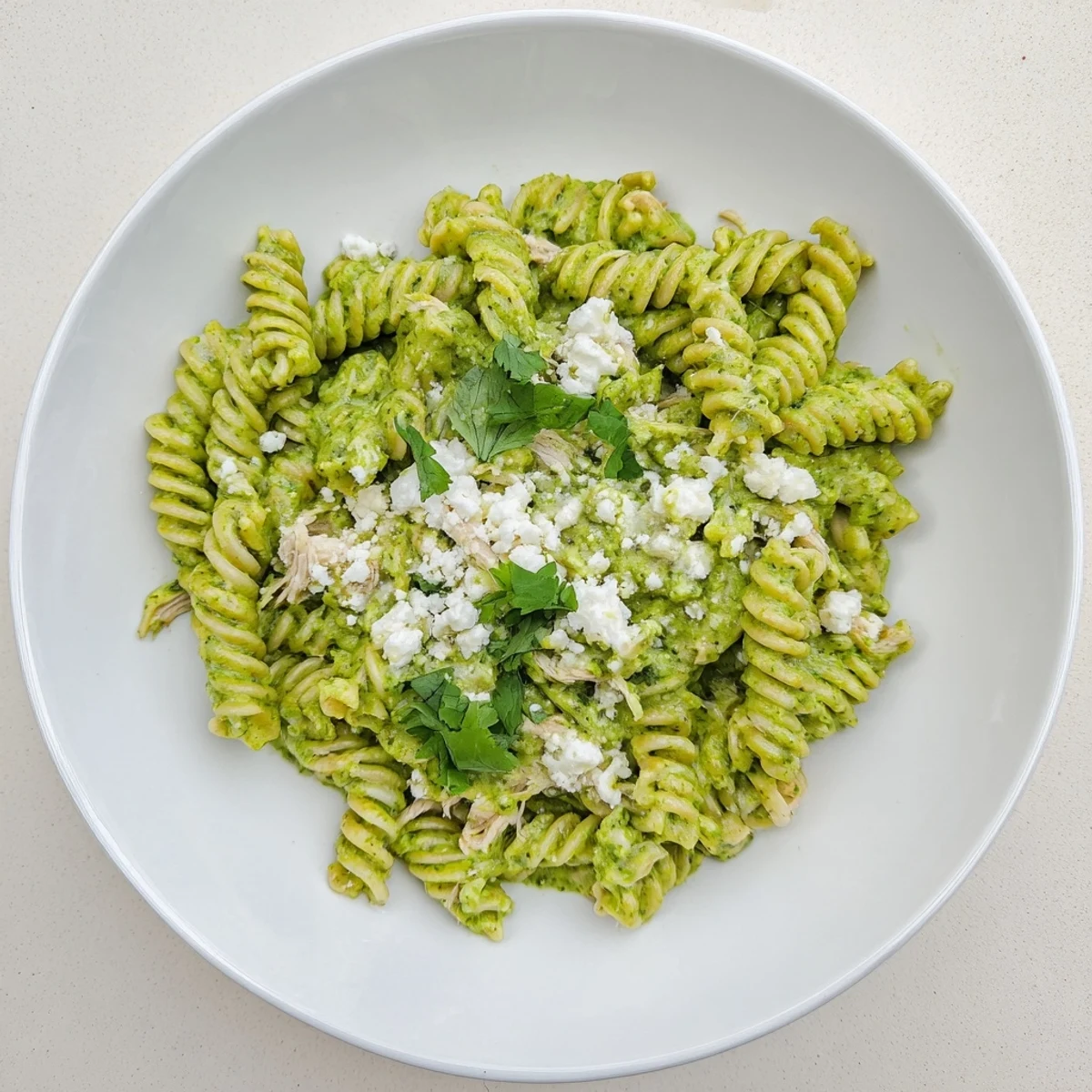 Creamy Cilantro Lime Chicken Pasta with rotini, shredded chicken, and a vibrant green yogurt sauce, garnished with crumbled cotija cheese and fresh lime wedges.  
